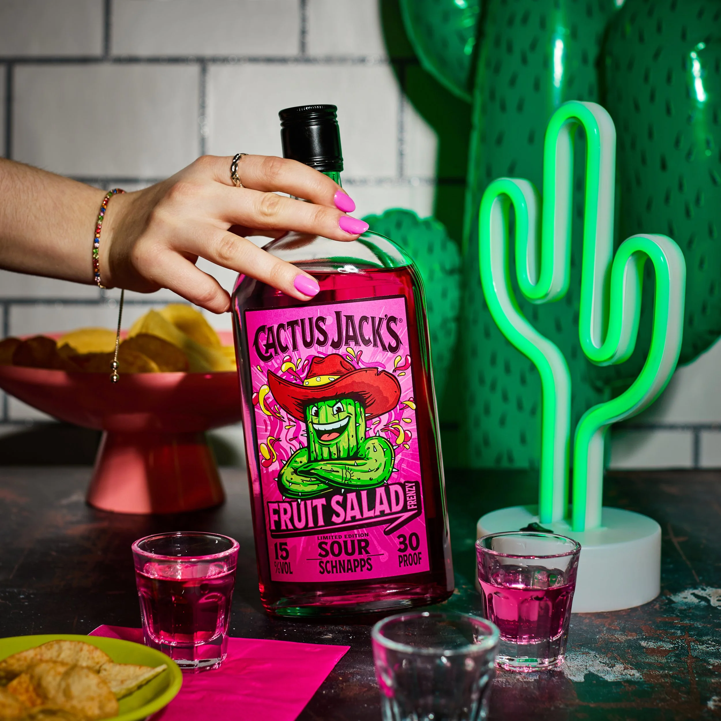 cactus jacks fruit salad flavour bottle  held by female with pink nails, with neon cactus sign, shot glasses and crisp snacks
